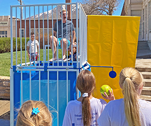 A teacher sits in a dunking booth while students line up to throw a ball at the target.